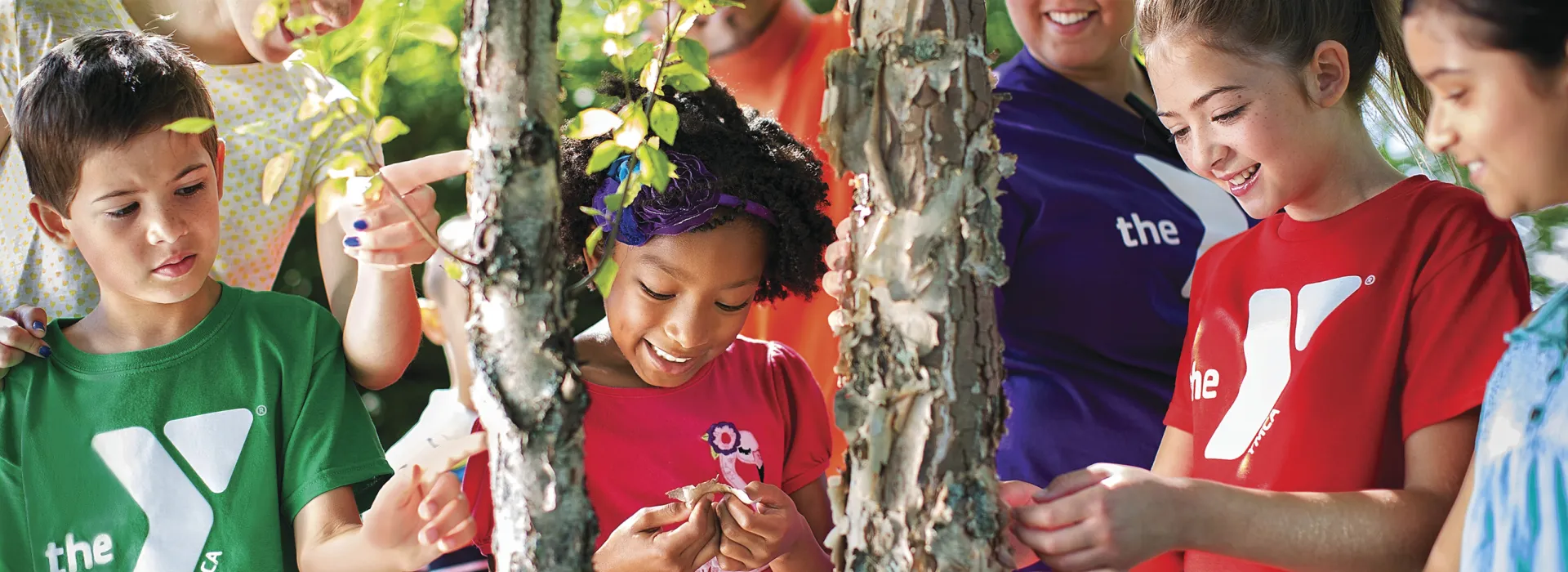 Children participating in an outdoor camp activity. They are huddled around a small tree studying its leaves.
