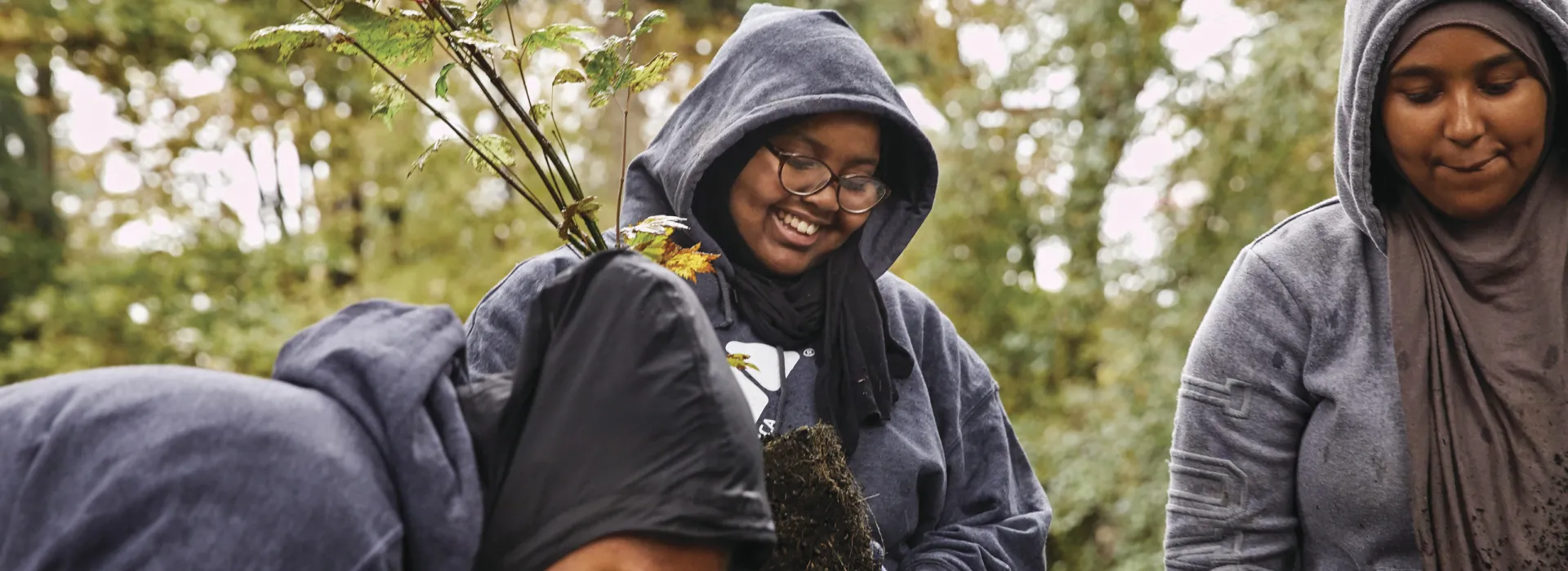 Three young adults huddled together. They are outside with tree saplings in their hands.